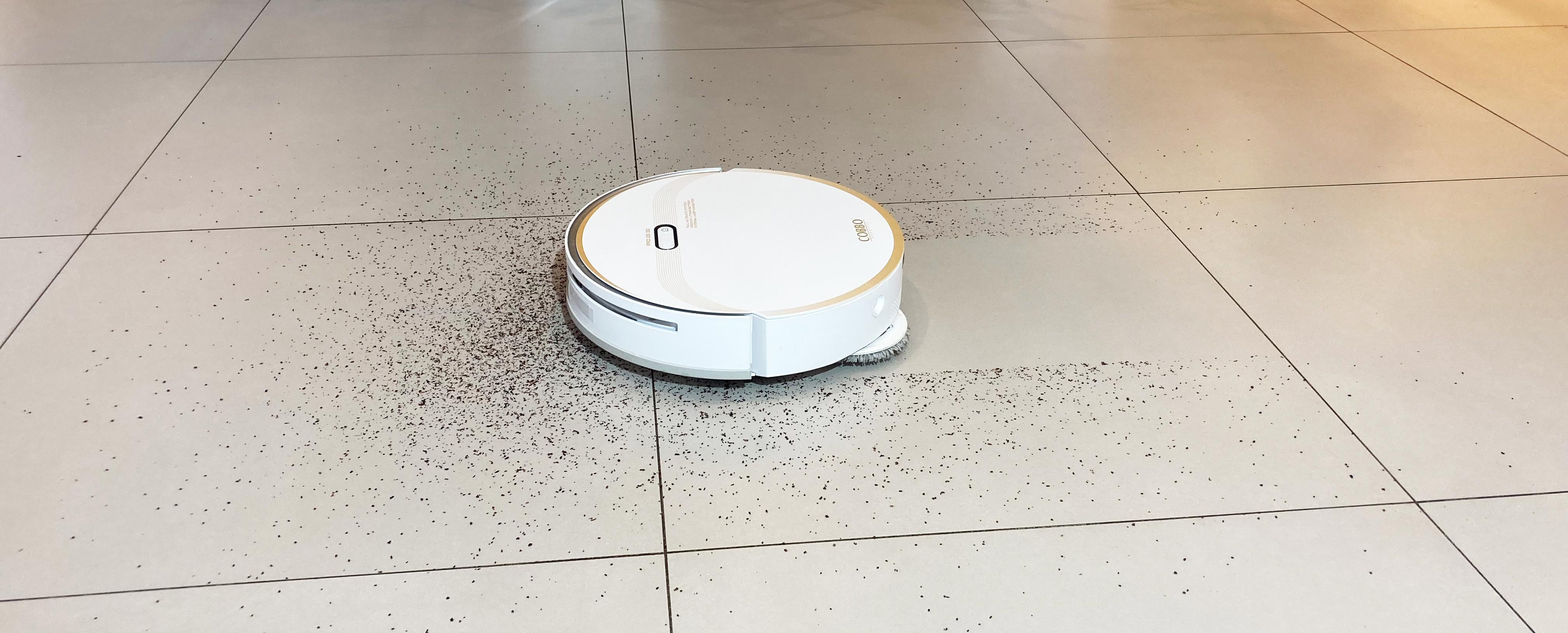 biały okrągły robot sprzątający jeżdżący po jasnych płytkach, na kt&oacute;rych jest rozsypana kawa